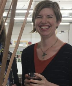 A young white woman with short brown hair holds a wine glass. She is smiling and standing next to willow rods in a large exhibition space with fluorescent tube lighting.