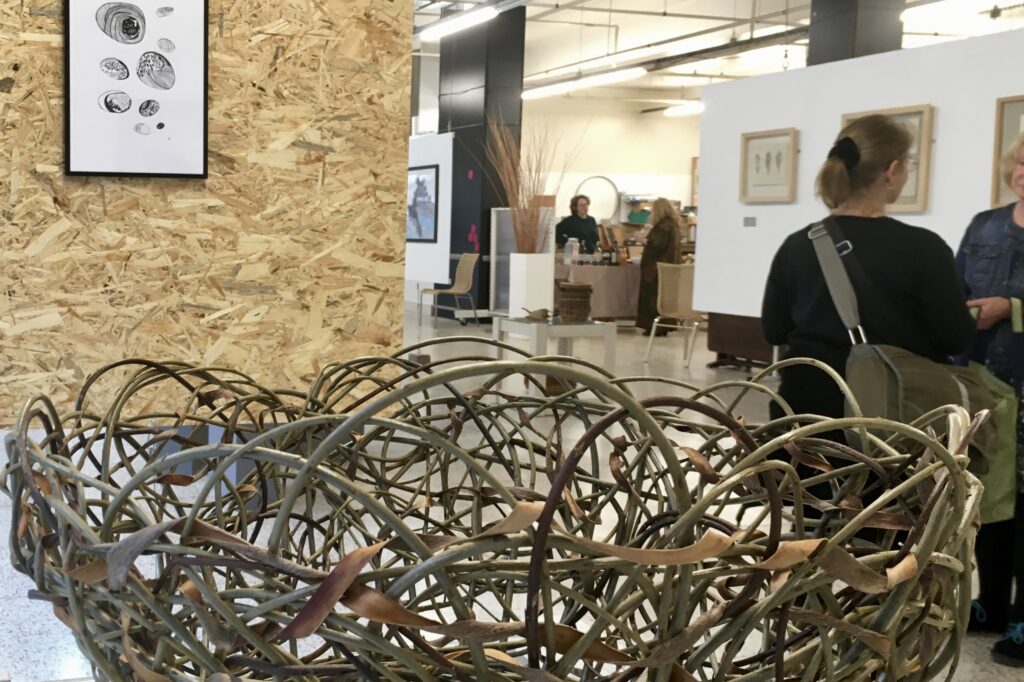 An exhibition installation in a large industrial space with a woven basket in the foreground, behind it a framed ink, drawing of pebbles displayed on a wooden board to the left, two women talking to the right.